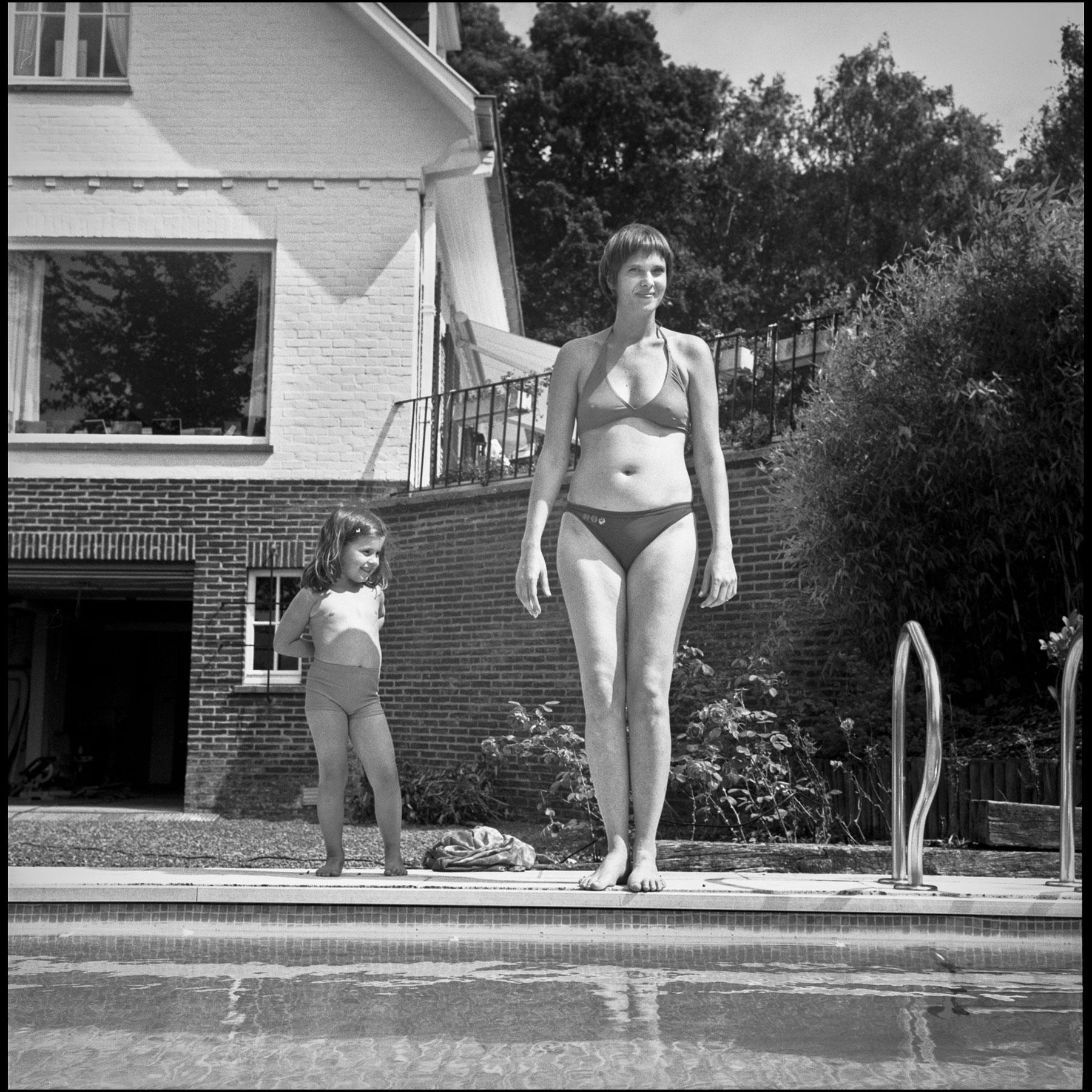 fillette-femme-maillot-de-bain-debout-bord-de-piscine-devant-maison-brique-noir-et-blanc